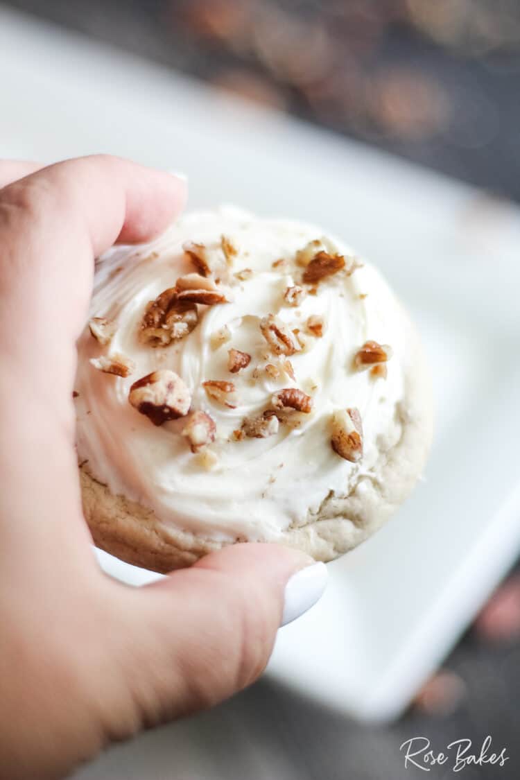 Manicured hand holding a butter pecan cookie with frosting and pecans on top.