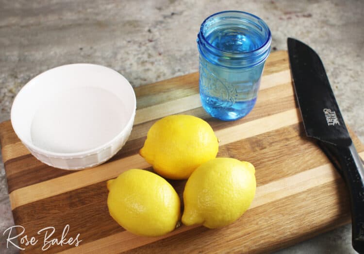 lemons, white bowl of sugar, blue cup of water and knife on a cutting board