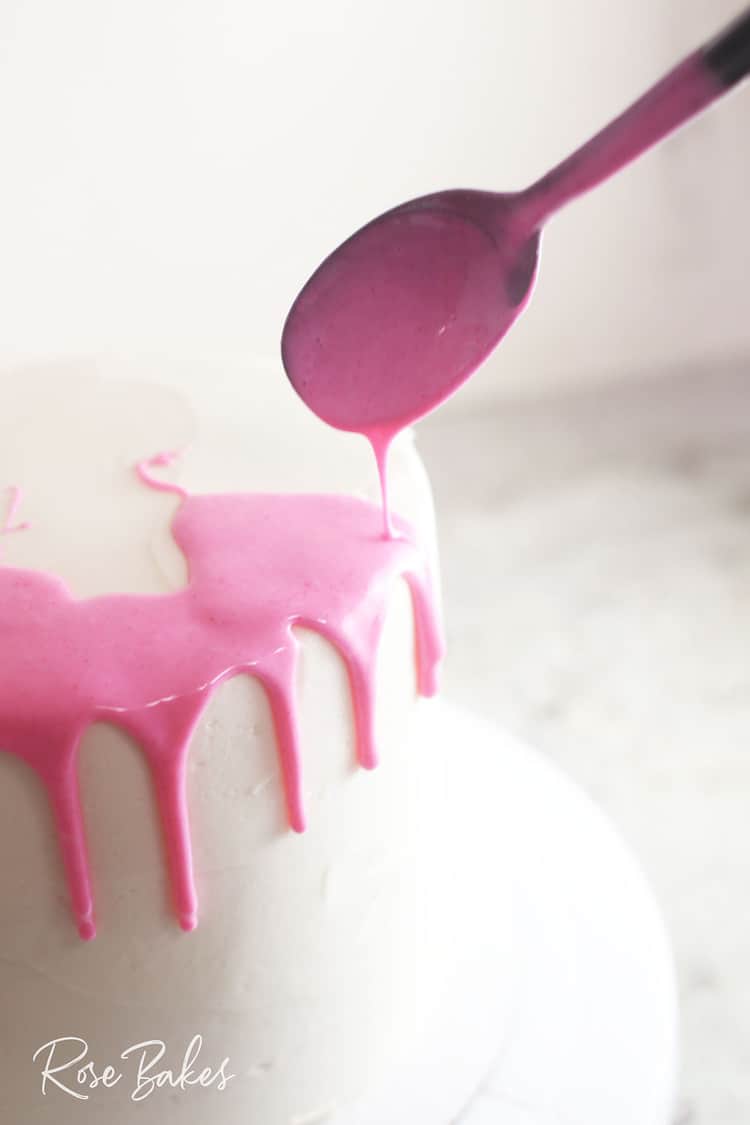 Pink canned frosting being spooned onto the edge of a chilled white cake to create a drip cake
