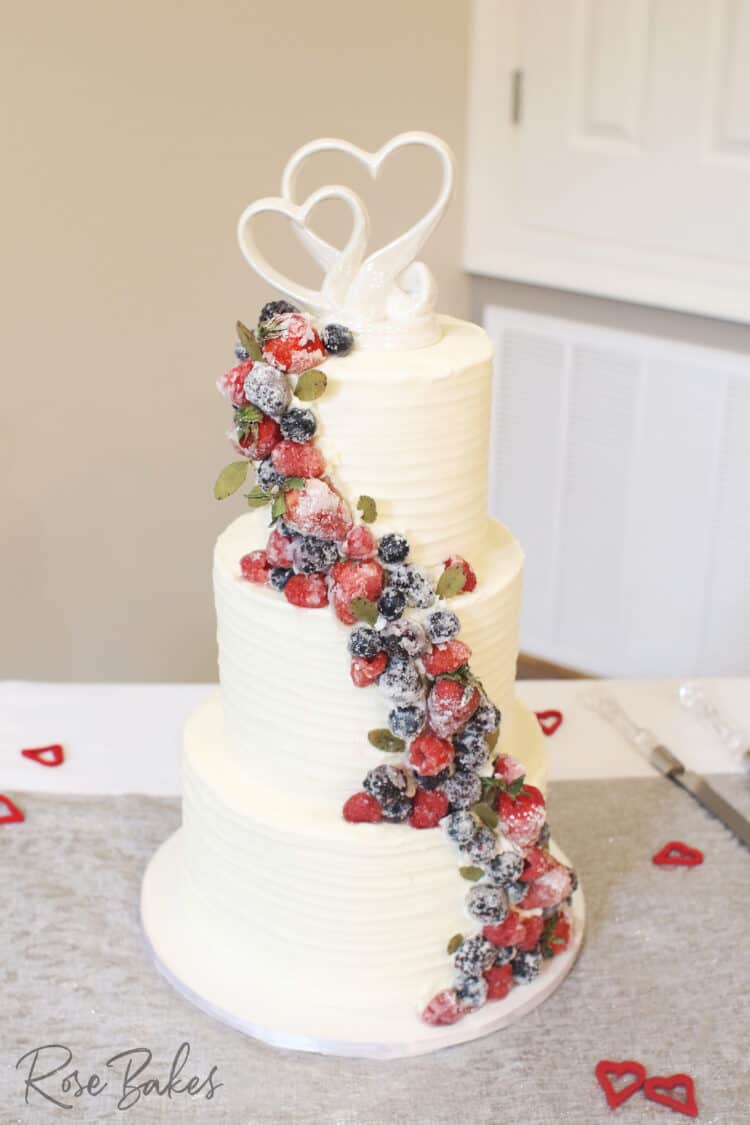 Three tiered wedding cake with textured buttercream and cascading sugared berries.  The topper is white interlocking hearts.
