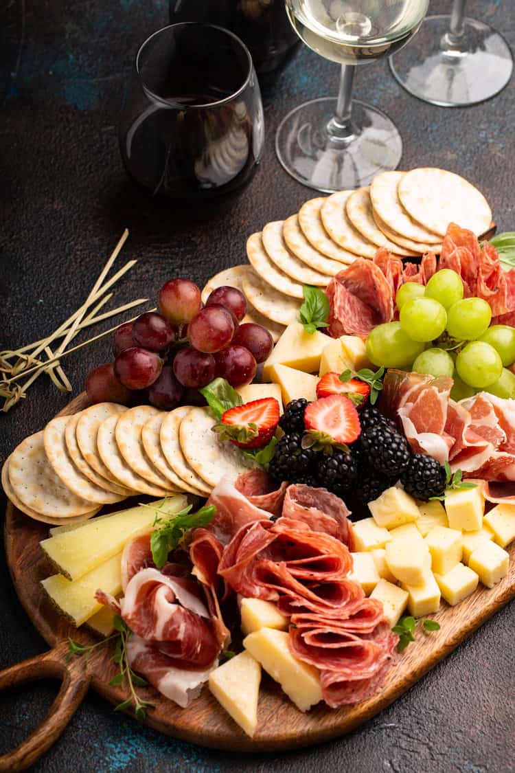 Charcuterie board filled with meats, cheeses and fruits with wine glasses next to it on a black counter