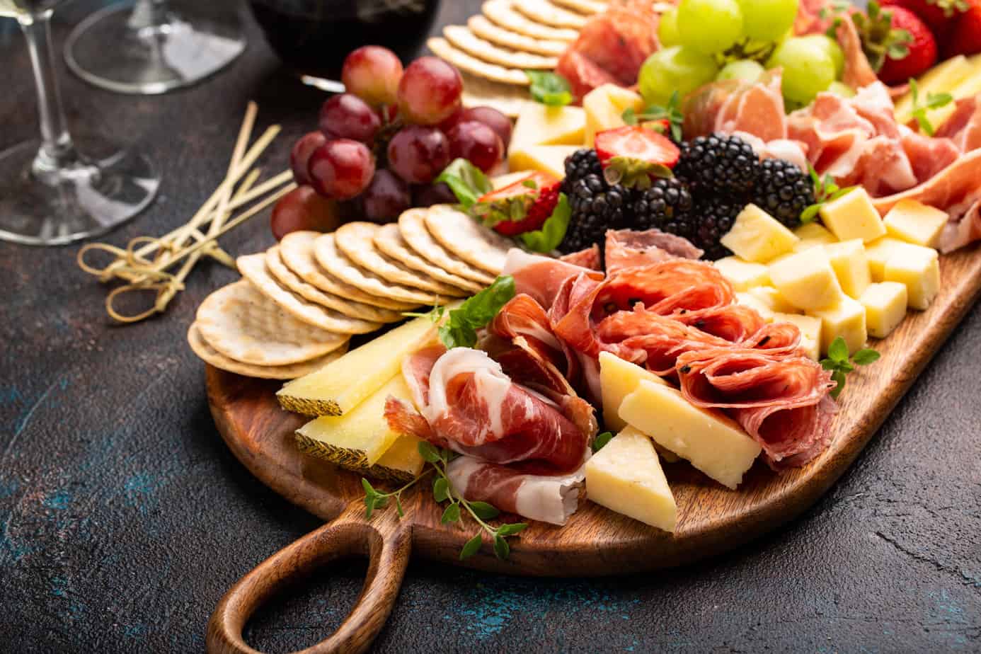 Charcuterie board filled with meats, cheeses and fruits with wine glasses next to it on a black counter 
