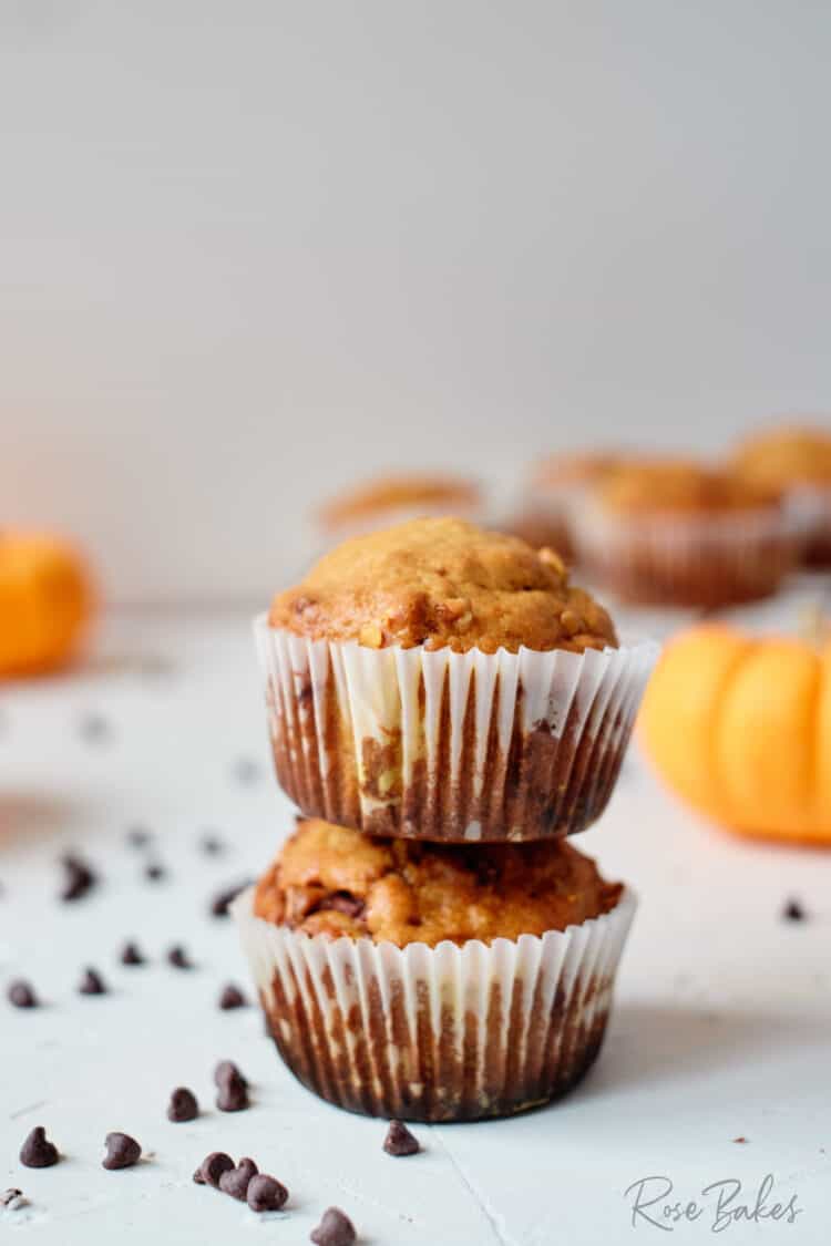 A couple of muffins stacked with chocolate chips scattered around on the table and mini orange pumpkins in the background.