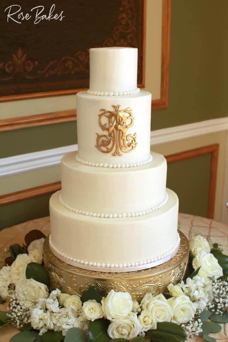 Gold monogram wedding cake on a gold stand surrounded by fresh flowers