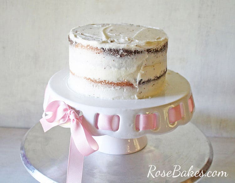 crumb coated cake on white cake stand with pink ribbon - waiting for messy ruffles buttercream