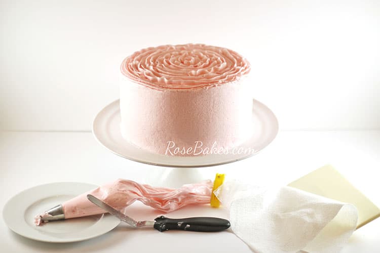 A light pink cake is displayed on a white cake stand. Below the cake is a piping bag filled with pink frosting, an offset spatula, paper towel and smoother.