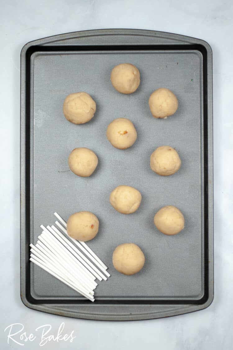 Cake pops rolled into balls on a baking sheet.  White pop sticks are in a stack at the bottom of the pan.