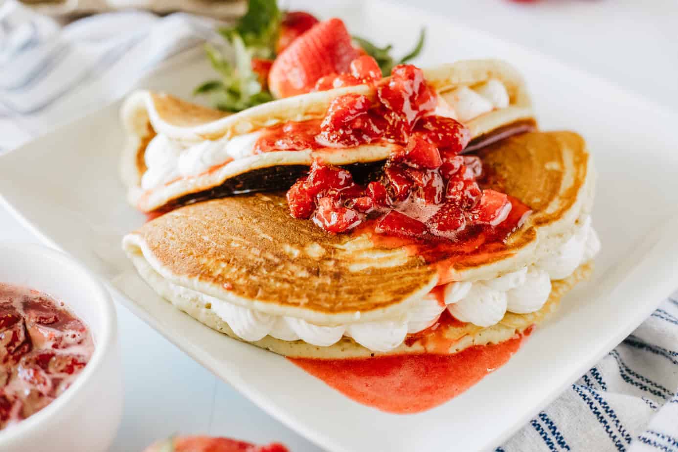 two buttermilk pancakes stuffed with no-bake cheesecake filling folded like tacos on a white plate with fresh strawberries and strawberry syrup on top