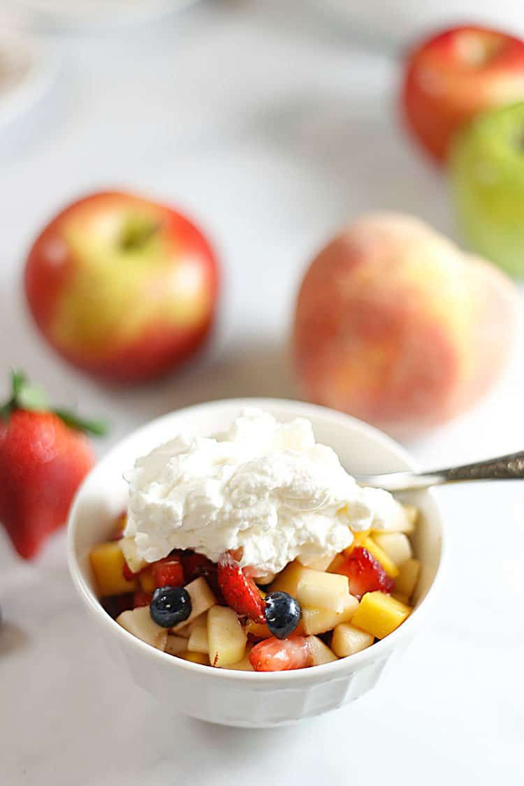 White bowl filled with fruit salad and topped with homemade whipped cream. Whole fresh fruit is in the background.