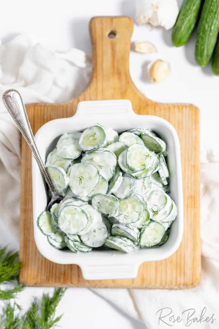 german cucumber salad tossed in a creamy white german dressing in a white square bowl sitting on a cutting board with a spoon