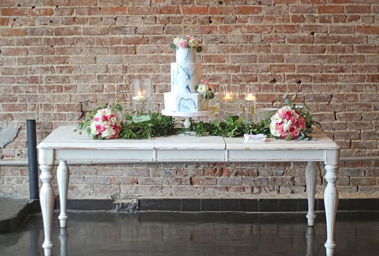 Marbled Fondant Wedding Cake with Navy, Gold and Pink on a white table with greenery, pink bouquets, and candles with brick background