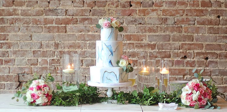 This Marbled Fondant Wedding Cake with Navy, Gold and Pink on table with greenery and pink bouquest and brick background