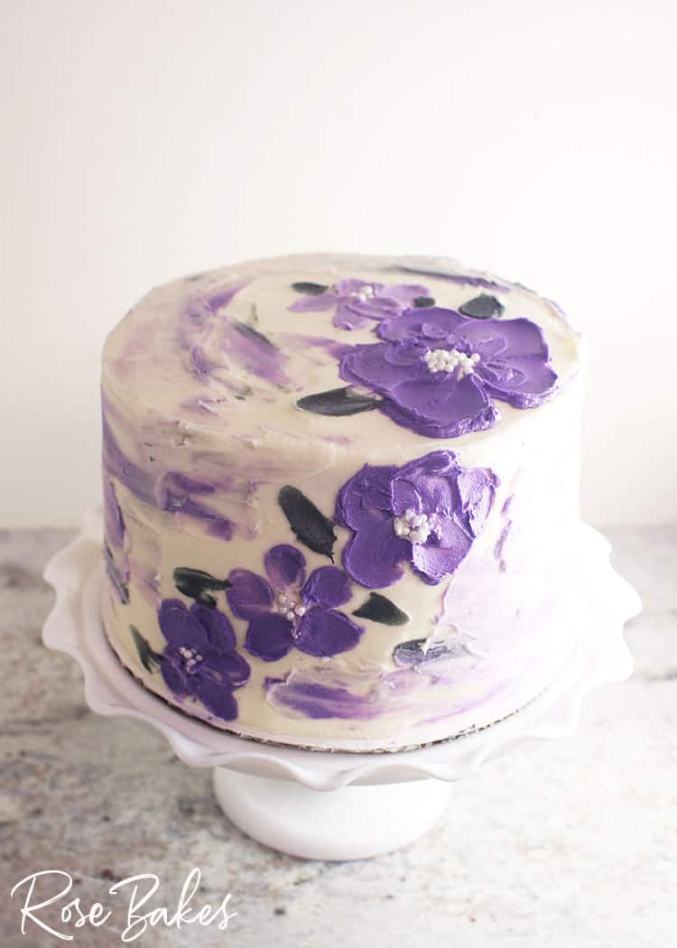 A white buttercream cake with purple watercolor streaks and purple flowers created with a palette knife and white sprinkles for the centers.  The cake is displayed on a white cake stand with a large ruffled edge.