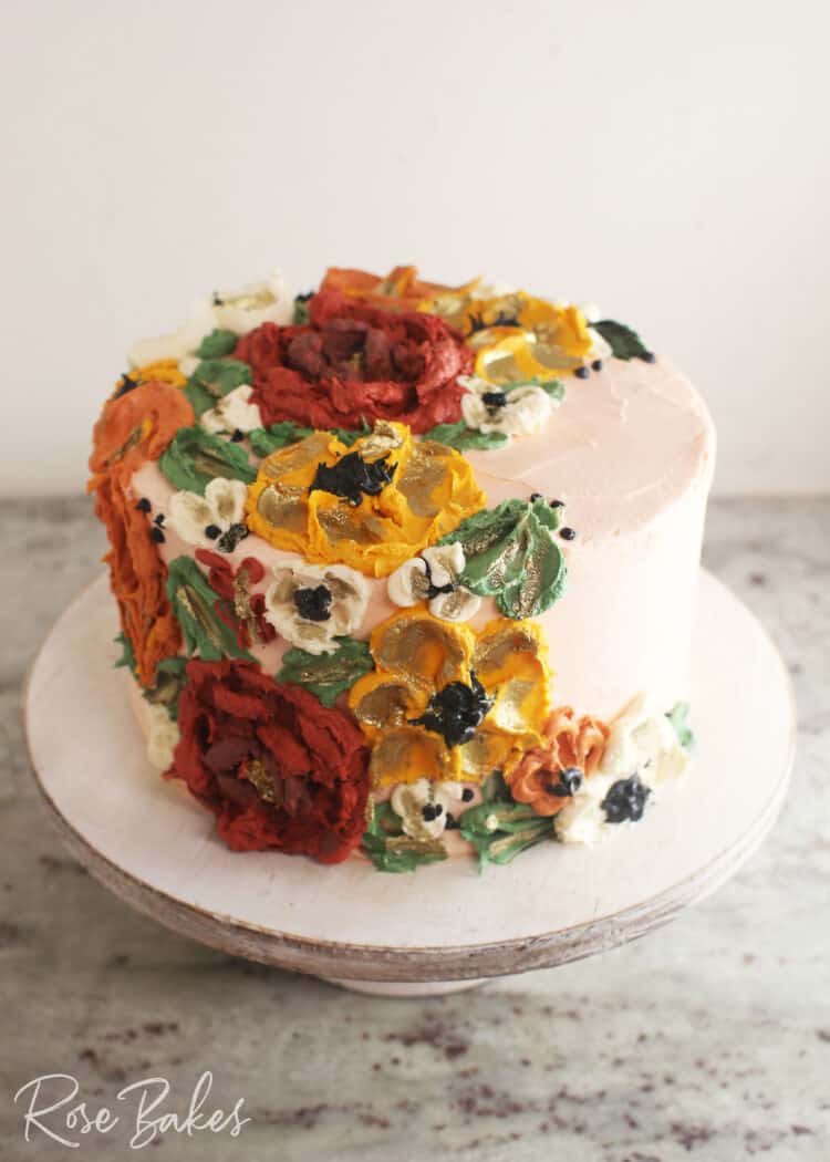 Cake frosted white with green leaves and red, yellow, and white buttercream flowers created with palette knives on one side of the top of the cake and cascading down the side of the cake.  
