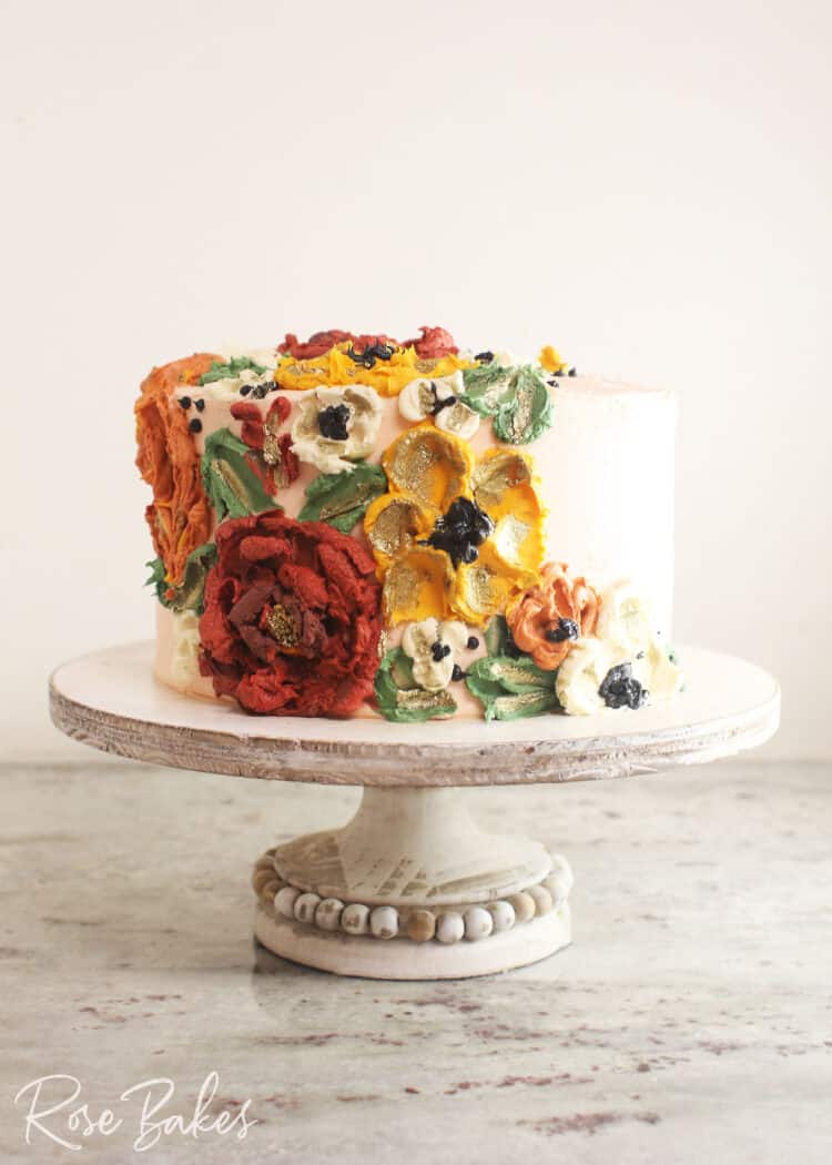 Front on view of a cake frosted white with green leaves and red, yellow, and white buttercream flowers created with palette knives on one side of the top of the cake and cascading down the side of the cake. The cake is displayed on a rustic wooden cake stand with a bead border around the base of the stand.