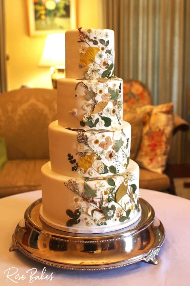 side view of pressed flowers wedding cake on silver stand on table with white tablecloth