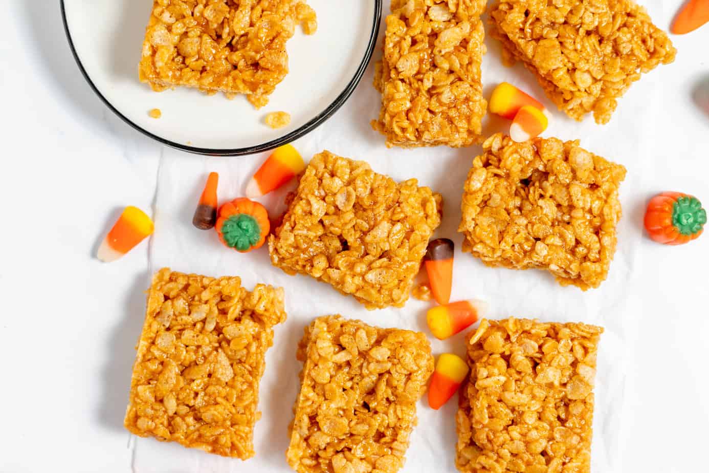 Pumpkin Rice Krispie Treats scattered on a white surface with two on a plate and some candy corn scattered near them