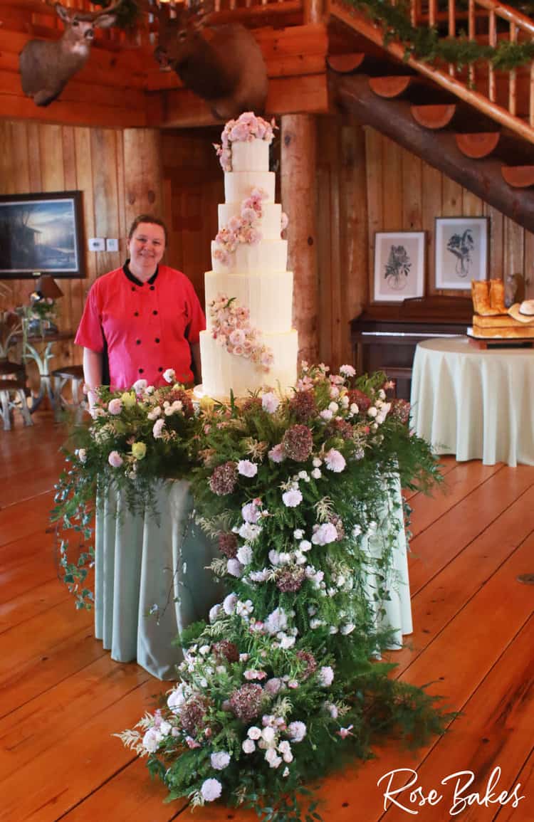 Rose standing next to Vertical Lines Buttercream Luxury Wedding Cake on a table filled with fresh flowers that are pink and white