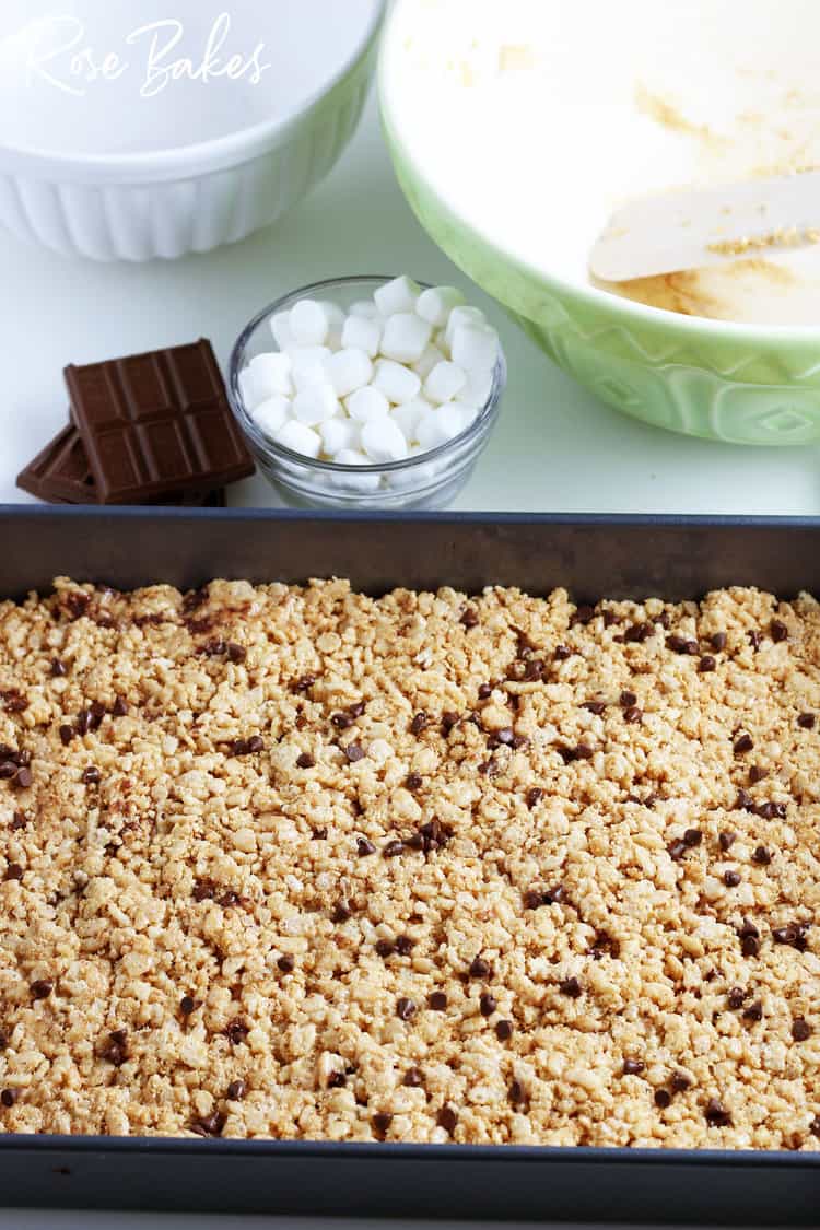 S'mores Rice Krispie Treats spread in baking dish to cool with small bowl of mini marshmallows and chocolate bars next to the pan.