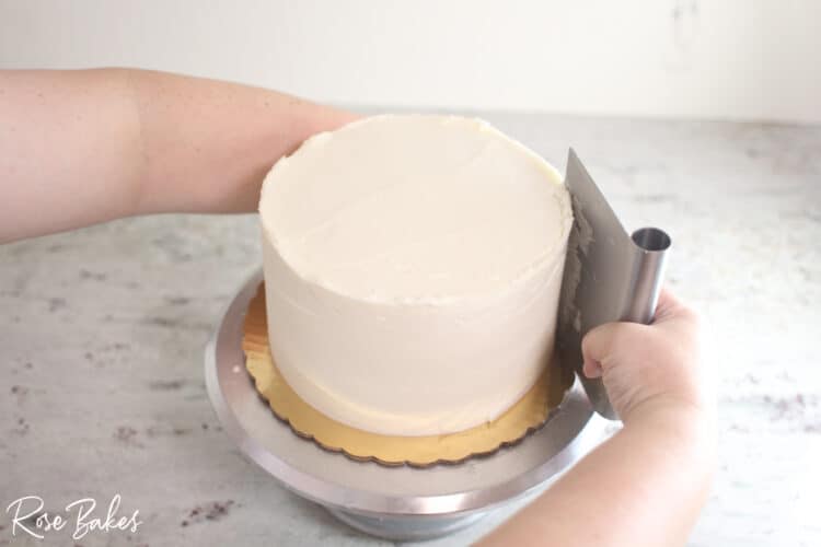 buttercream cake on turntable being smoothed with benchscraper for sharp edges