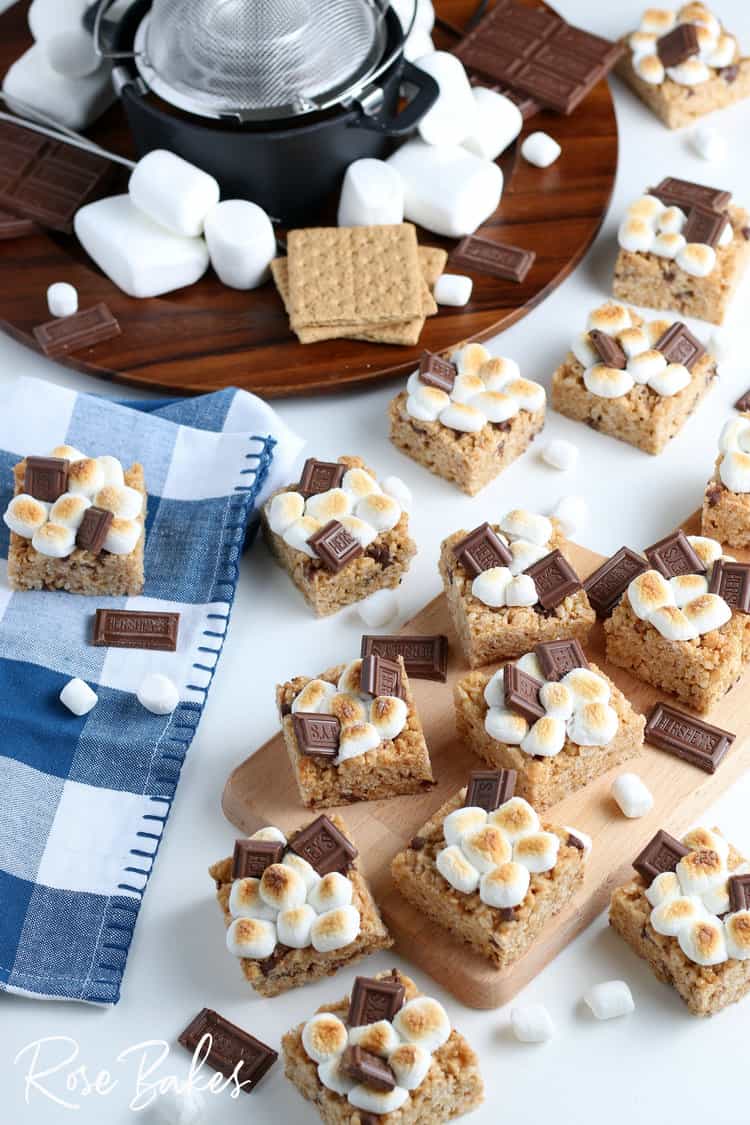 Smores Rice Krispie Treats cut into squares and spread on a cutting board and checkered kitchen towel. They are topped with toasted mini marshmallows and a piece of chocolate.