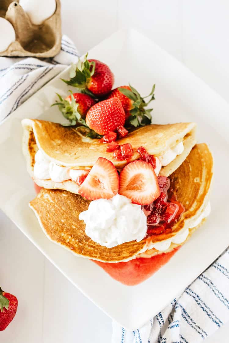 Strawberry Cheesecake Pancakes on a white plate. Pancakes folded over cheesecake filling with strawberry syrup, whipped cream and fresh strawberries