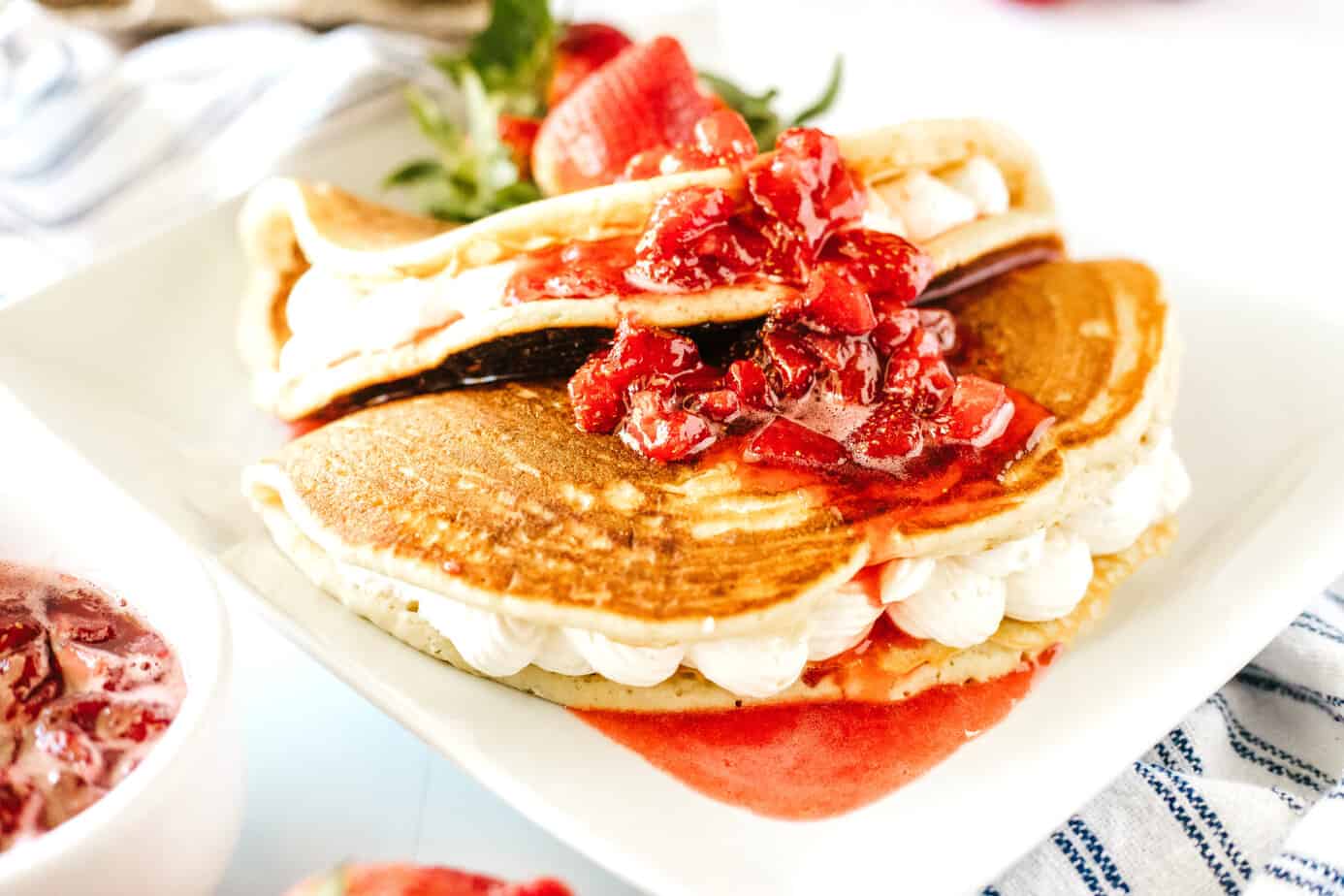 Strawberry Cheesecake Pancakes on a white plate. Pancakes folded over cheesecake filling with strawberry syrup, whipped cream and fresh strawberries