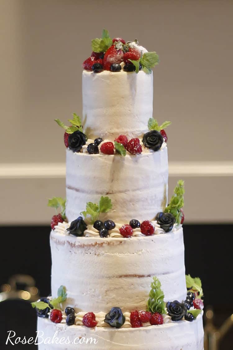 4 tier rustic buttercream cake with sugared berries, fresh mint, and black sugar roses around the top of each tier.