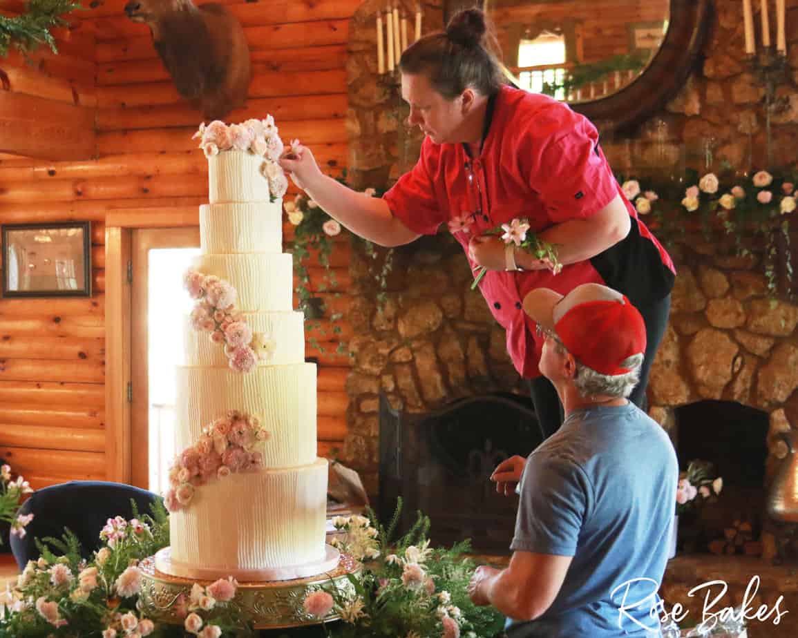 Rose standing on a stool adding flowers to a tall vertical lines in buttercream wedding cake