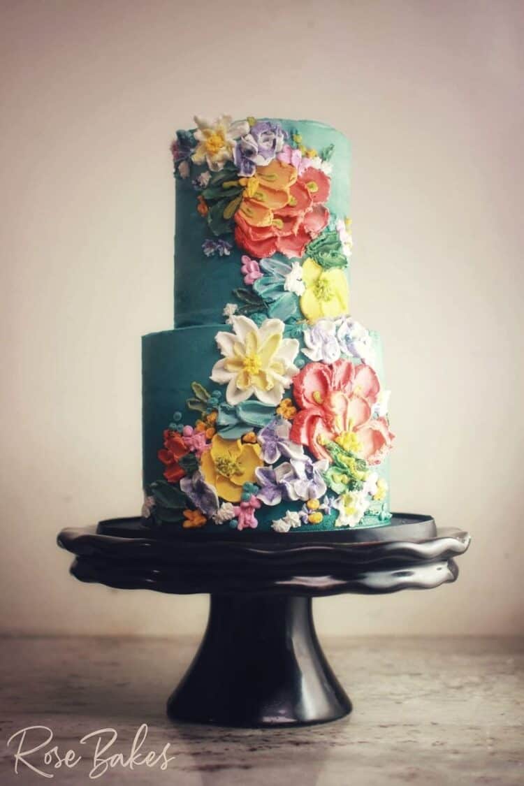 3 tier deep green buttercream icing with buttercream flowers painted on in multi fall colors on black cake stand 