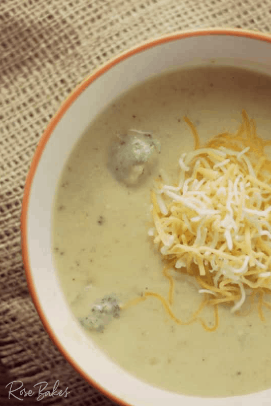 Close up of Broccoli Cheese Soup topped with shredded cheese in an orange rimmed bowl on a burlap placemat.