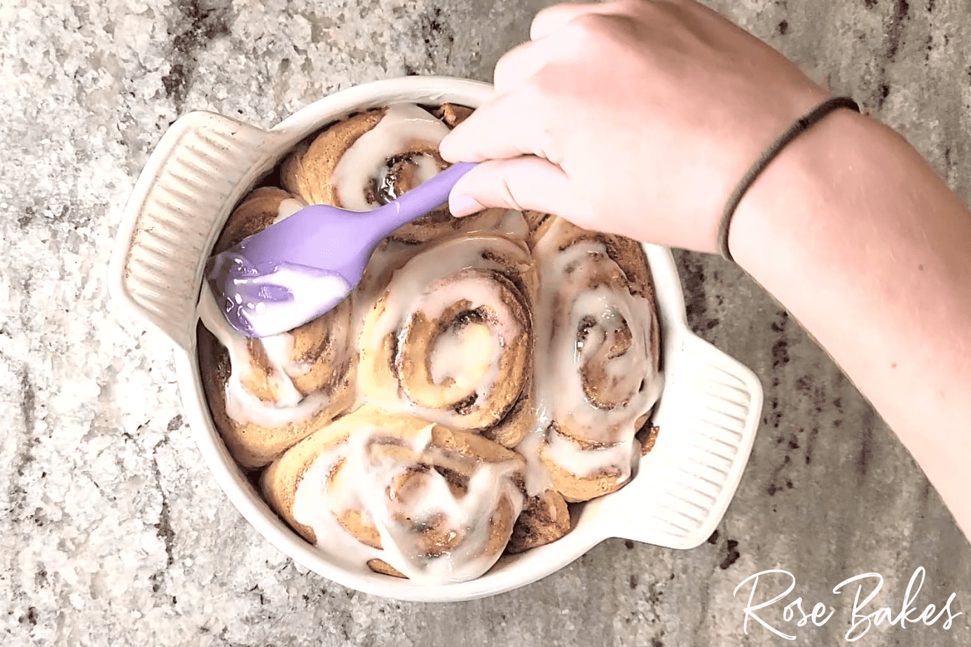 Cream Cheese Icing being spread on top of the baked cinnamon rolls with a purple spatula.